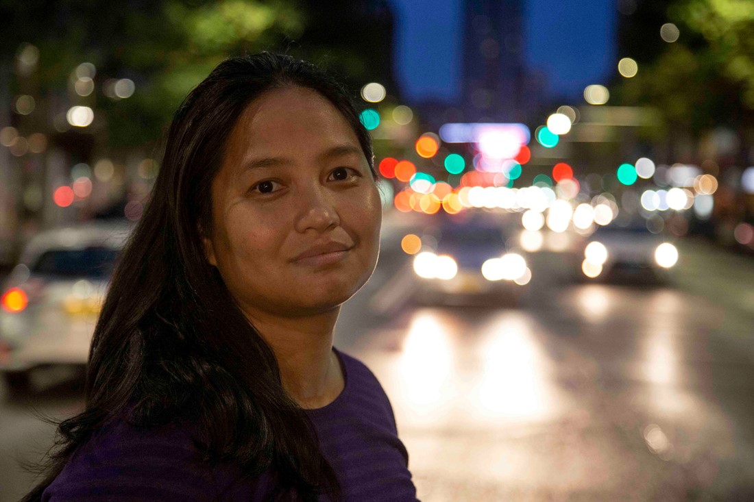Portrait shot of Jai, smiling, in the evening. Behind her, the lights of a bustling city.