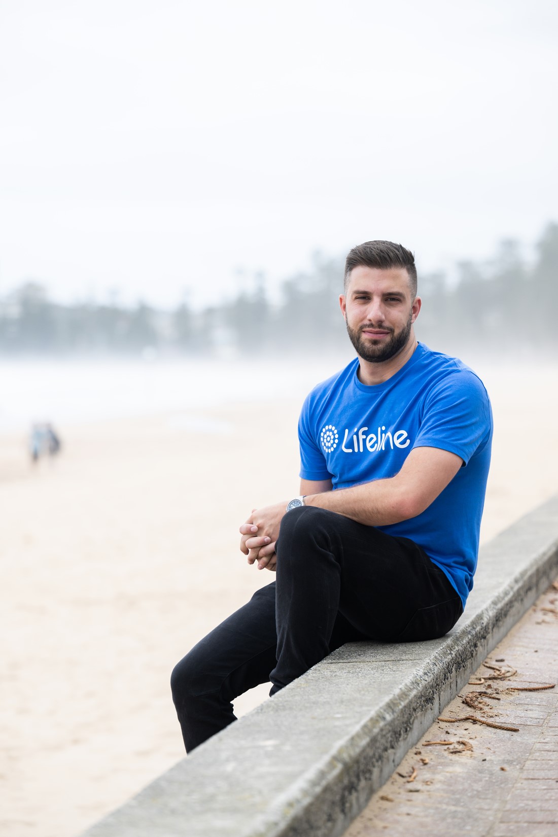Michael sitting on the steps of Manly Beach on an overcast day, wearing a blue Lifeline t-shirt.