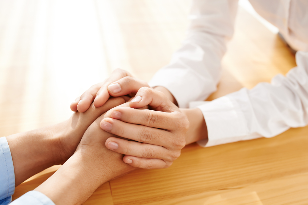 Two hands, one gently covering the other, are clasped together on a light wooden table. The image suggests comfort, support, and human connection, reflecting empathy and care.