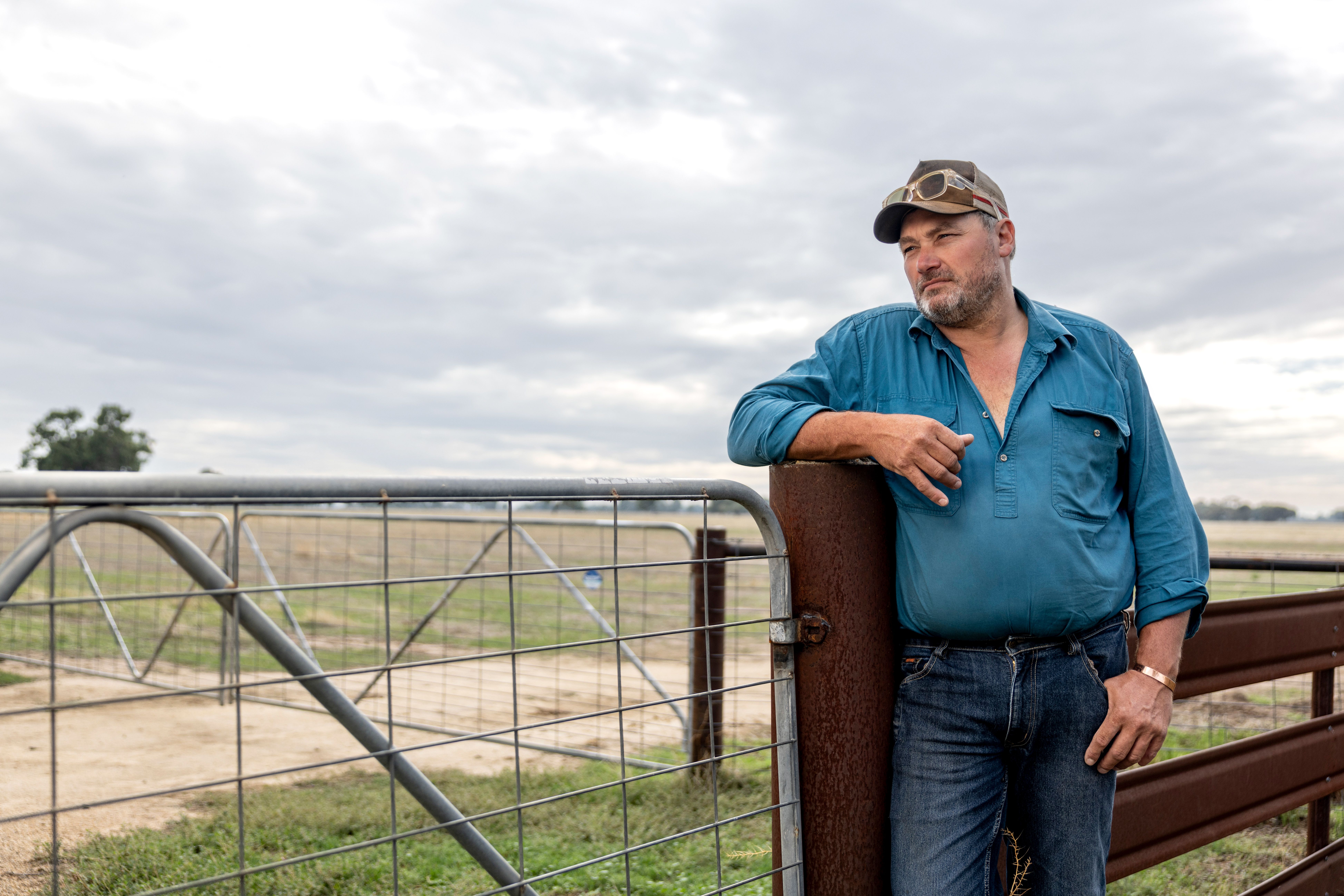 A middle aged Australian man in a rural setting, one arm learning on a gate, looking off into the distance.