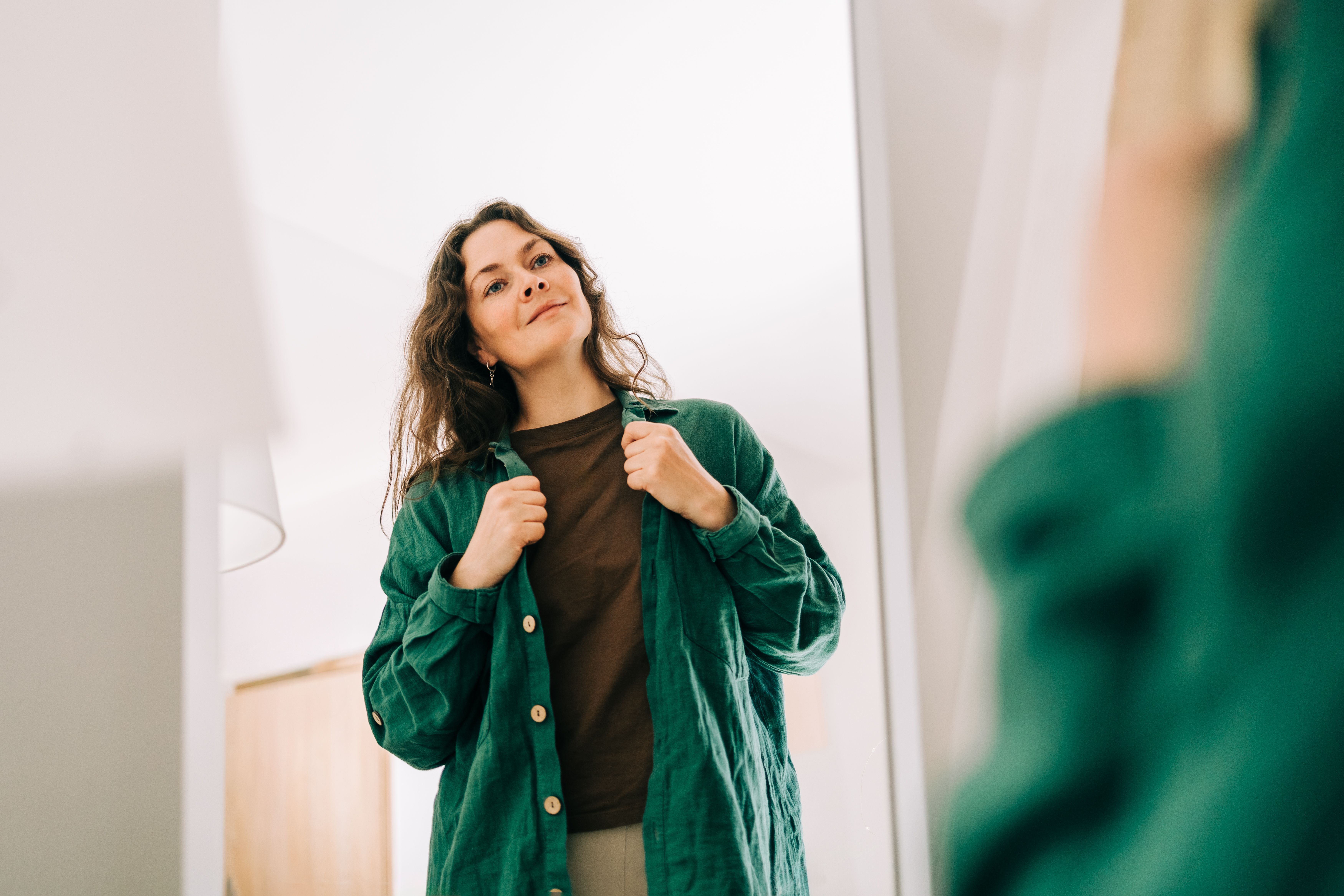 A woman wearing a green overshirt looks at herself in a mirror while adjusting her collar.