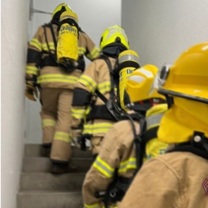 Firefighters in full protective gear, including yellow helmets and breathing apparatus, ascend a stairwell. The image highlights a physical challenge with text overlay: "28 floors, 25kg of kit".