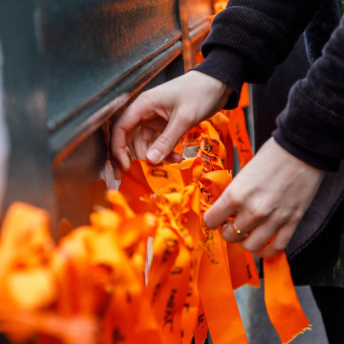 A person's hands gently arranging or tying numerous bright orange ribbons, some bearing faint handwritten messages, onto a dark metal structure. The orange ribbons are a symbol of suicide prevention awareness and remembrance.