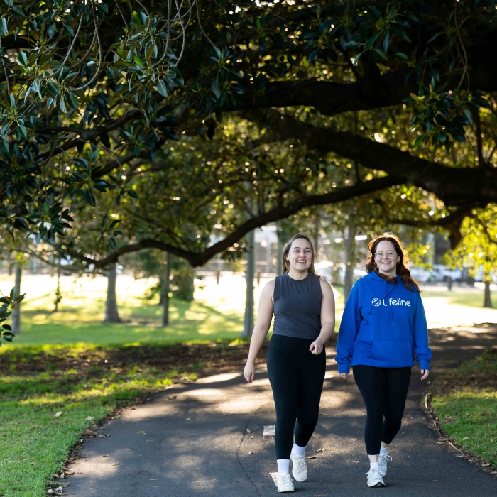 Two young women are smiling and walking together on a paved path in a sunny park. The woman on the right is wearing a bright blue hoodie with the white "Lifeline" logo prominently displayed on the front. This image promotes positive well-being and connection.
