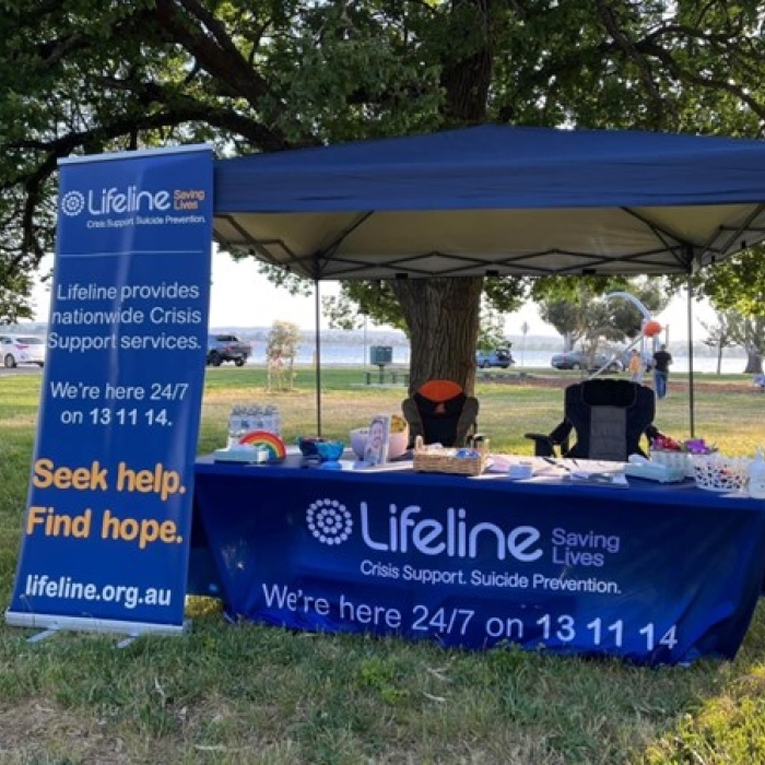 A Lifeline outreach tent is set up in a park, featuring a blue canopy and a table covered with a branded tablecloth. A prominent vertical banner next to the tent displays the Lifeline logo and key messages: "Crisis Support. Suicide Prevention. Lifeline provides nationwide Crisis Support services. We're here 24/7 on 13 11 14. Seek help. Find hope. lifeline.org.au". The table is laden with informational materials and small support items, highlighting Lifeline's presence and availability for crisis support.