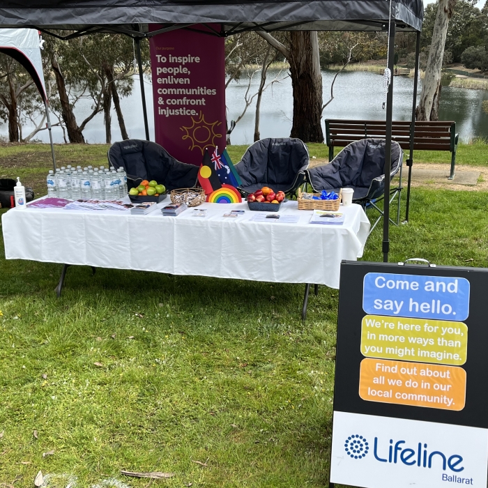 A Lifeline Ballarat outreach tent is set up in a grassy park by a lake. Under a black canopy, a white-covered table offers water bottles, fruit, and information. A purple banner displays the text: "Our purpose: To inspire people, enliven communities & confront injustice." A freestanding A-frame sign invites, "Come and say hello. We're here for you, in more ways than you might imagine. Find out about all we do in our local community. Lifeline Ballarat."