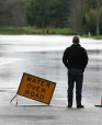 A man standing on a road in front of floodwaters, looking away from the camera. A sign reads "WATER OVER ROAD" to his left, to warn drivers.