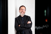 Dr Tim Sharp smiling with his arms crossed, wearing glasses and a black suit.