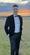 Mark smiling in a black suit and bow tie, standing outdoors at sunset.