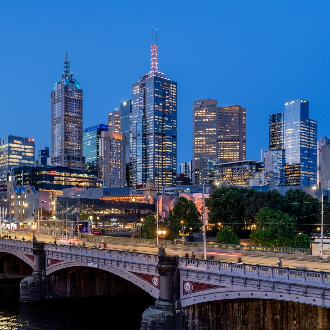 Melbourne city skyline at dusk, featuring Princes Bridge spanning the Yarra River in the foreground, with blurred light trails from traffic. Illuminated skyscrapers, including the Eureka Tower and Rialto Towers, rise in the background. The dome of Flinders Street Station and the spires of St Paul's Cathedral are visible on the left, reflecting a vibrant urban landscape.