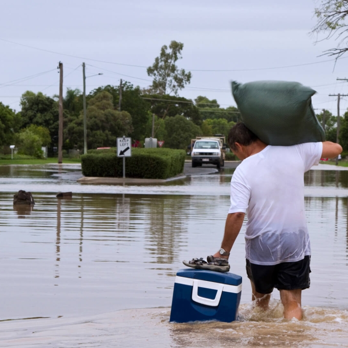 A man wades through knee-deep floodwater in a residential area, carrying a large green bag over his shoulder while pulling a blue cooler with one hand. He wears a white t-shirt, black shorts, and sandals, navigating the submerged street where trees, utility poles, and road signs are partially underwater. A vehicle is visible in the distance on higher ground. The scene depicts the challenges faced during severe flooding.