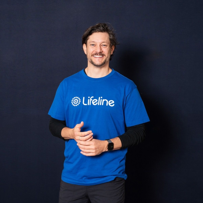 Rob Mills, an Australian singer and actor, smiles while wearing a blue Lifeline t-shirt over a black long-sleeved shirt. He has dark, wavy hair, a beard, and a watch on his left wrist. His hands are clasped together in front of him, and he stands against a dark blue background.