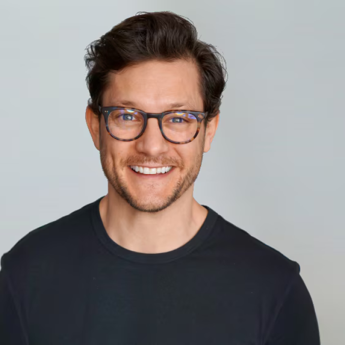 Rob Mills, a smiling man with dark hair and tortoise-shell glasses, wearing a black t-shirt against a light grey background.
