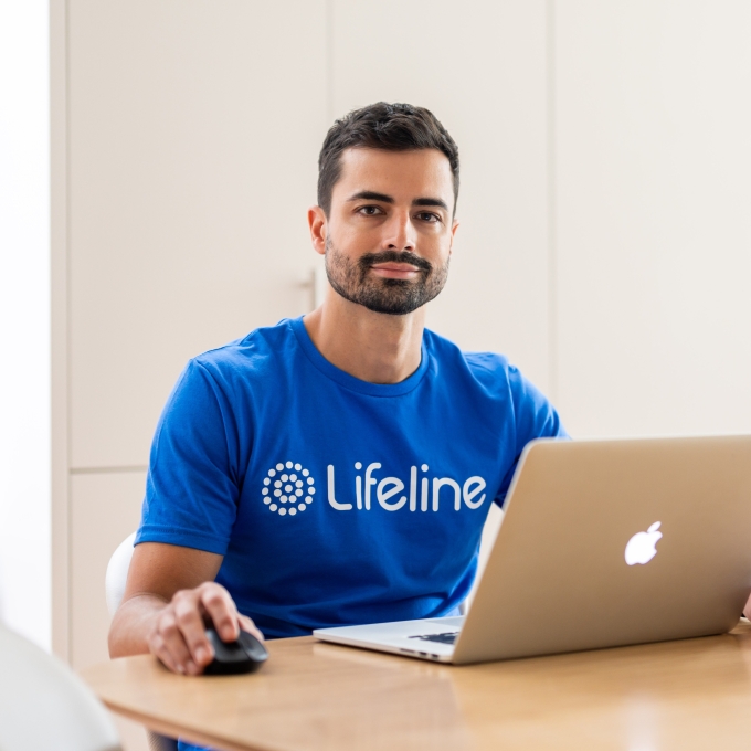 A man with dark hair and a beard, wearing a blue Lifeline t-shirt, sits at a light wooden table. He is holding a computer mouse with his right hand and has a silver laptop open in front of him. He is looking directly at the camera with a calm, friendly expression. The background is a bright, modern kitchen with light-coloured cupboards.