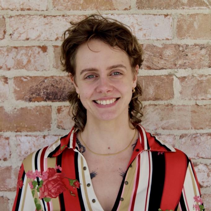 Jay Gardener, wearing a striped button down shirt, standing in front of a brick wall.