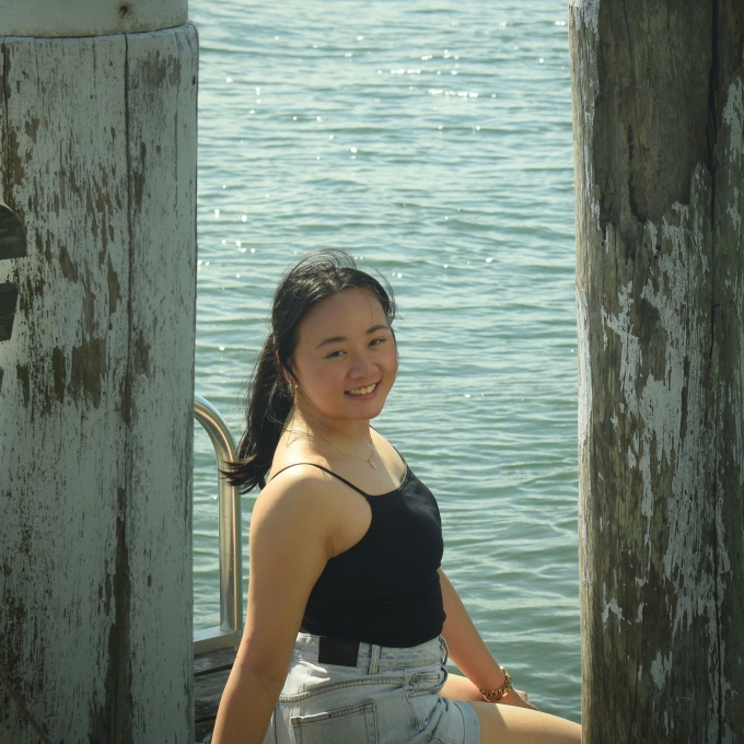 Yasmin smiling while sitting on a wharf beside the water, framed between two weathered timber posts.