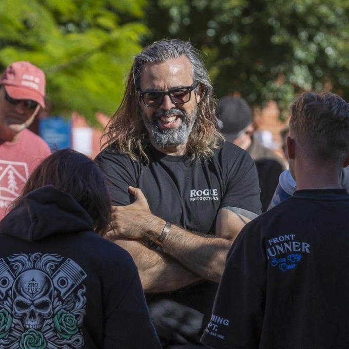 Stephen smiles while talking with a group of people outdoors, wearing sunglasses and a black Rogue Motorcycles T-shirt.