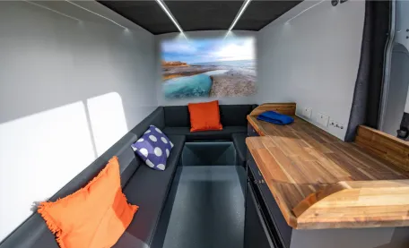 Modern camper van interior with a large L-shaped dark gray bench seating area, accented with two orange and one blue polka-dot cushion. Above the far end of the bench is a bright landscape image of a rocky shoreline and calm turquoise water. To the right, a wooden countertop with a dark blue towel sits above built-in cabinets. The walls are white, the ceiling is dark gray with linear LED lighting, and the floor is dark gray. Sunlight streams in from an unseen window on the left.